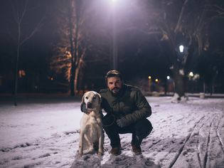 一位30多岁的白人男子蹲在一只金发的拉布拉多猎犬旁边。他穿着一件绿色的冬衣，手套，牛仔裤和棕色的鞋子。地上有雪，现在是晚上。